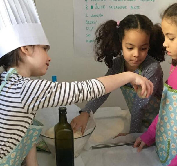 S kuharsko kapo pokrita deklica, ki ostalim otrokom prikazuje pripravljen cmok.