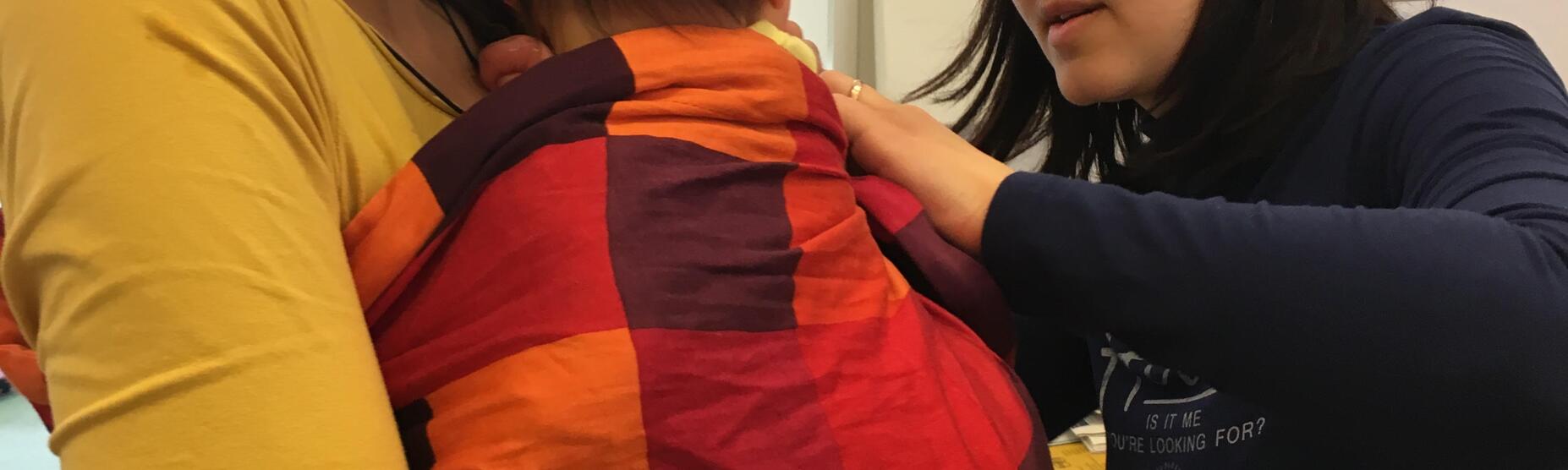 Mamica popravlja nosilno ruto drugi mamici z dojenčkom v ruti.