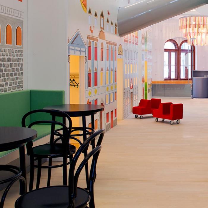 A photo of the interior of the playroom with a playfully painted wall, red armchairs on wheels and a table with chairs.